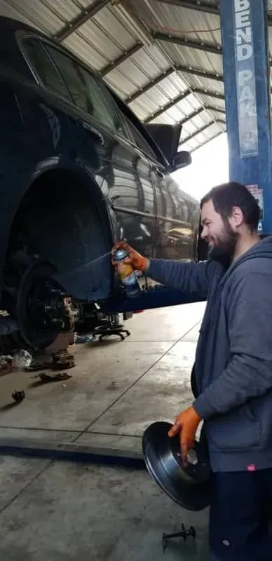 A man in a garage sprays a car part while holding another part. He wears gloves and a grey jacket.